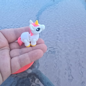 May include: A small, white unicorn figurine with a pink mane and tail, a yellow horn, and yellow hooves. The unicorn has a black eye and is held in a hand.
