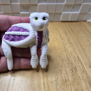 May include: A 3D-printed white turtle figurine with a purple shell design. The turtle has a detailed textured surface, green eyes, and a purple neck. The figurine is held in a hand, showcasing its size and intricate details.