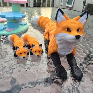 May include: An orange and white articulated fox toy with black legs and ears. Two smaller fox keychains are also present. The fox toys are on a glass table with a colorful, abstract structure in the background.