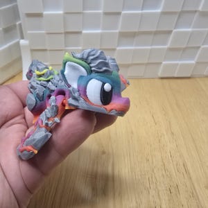 May include: A colorful, 3D-printed pony figurine with a unique rock-like texture. The pony features a rainbow-colored mane and tail, with a gray and stone-like body. The figurine is held in a hand, showcasing its intricate design and vibrant colors.