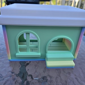 May include: A pastel-colored miniature house with a white roof and arched windows. The house features a mint green body with rainbow-colored edges and a small set of steps leading to an open doorway. The overall design is whimsical and playful.