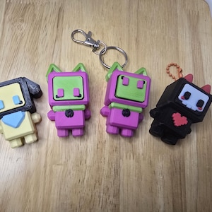 May include: A collection of colorful, geometric character keychains. The keychains feature various designs, including a yellow figure with a blue scarf, two pink figures with green accents, and a black figure with a red heart.