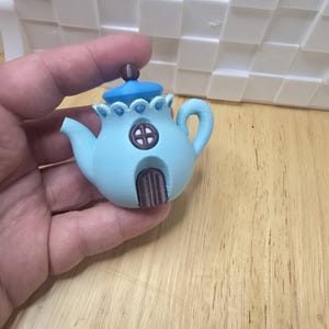 May include: A light blue teapot-shaped house with a brown door and window. The teapot has a blue lid and a brown knob. The handle and spout are curved. The teapot is held in a hand.