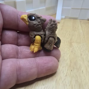 May include: A small, brown and yellow bird figurine with a detailed, textured surface. The bird has a yellow beak, talons, and jointed legs. The figurine is held in a person's hand.