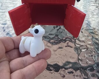 May include: A red and black barn with open doors and a white cross-shaped design. A small white goat figurine with black eyes and small horns is in front of the barn. The barn is on a glass table.