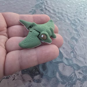 May include: A small, green, abstract sculpture with a textured surface and a light brown eye-like detail. The sculpture has a unique, organic shape and appears to be made of a solid material. The object is held in a hand.