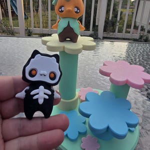 May include: A whimsical display featuring a pumpkin-headed figure with a green collar and brown body, perched atop a flower-shaped platform. A black cat figure with a skeleton design is held in front. The display has a pastel color scheme.