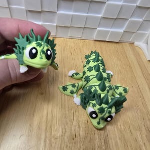 May include: Two green and white lizard figurines. One is held by a hand, the other is lying down. The lizards have large black eyes, white feet, and green leafy patterns. The background is a wooden surface.