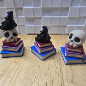 May include: Three miniature sculptures: a black bird-like figure and two skulls, each atop a stack of three books. The books are blue, purple, and red. The background is a white, textured wall.