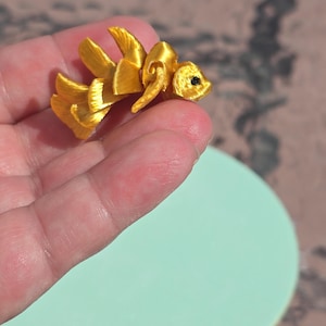 May include: A small, golden-yellow fish figurine with detailed fins and a small black eye. The figurine is held in a person's hand against a light green background. The fish appears to be made of a 3D-printed material.
