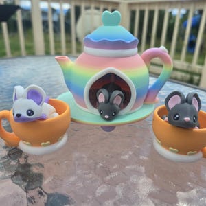 May include: A whimsical ceramic tea set featuring a rainbow-colored teapot with a heart-shaped lid and a mouse figurine inside. Two orange teacups with white bases each hold a mouse figurine. The set is on a glass table.