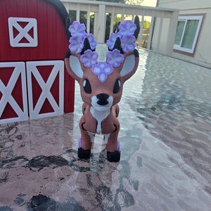 May include: A brown and white deer figurine with purple flower accents on its antlers. The deer is standing in front of a red barn with white doors and trim. The figurine is on a glass table.