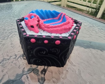 May include: A black hexagonal box with a pink and blue dragon sculpture on top. The dragon is curled up on a bed of white stones inside the box. The box has pink accents and decorative carvings.