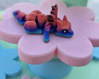 May include: A colorful, 3D-printed dragon figurine in shades of red, orange, blue, and purple rests on a pink flower-shaped platform. The platform sits atop a light blue base, with other flower-shaped elements in the background. The overall theme is whimsical and decorative.