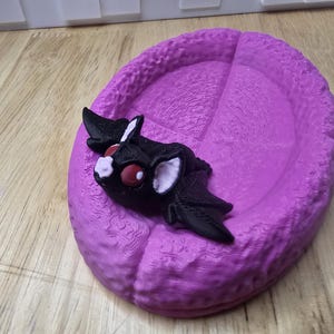 May include: A 3D-printed, purple, oval-shaped tray with a textured surface. A black bat-like creature with white and red accents rests on the tray. The bat has large ears and wings.