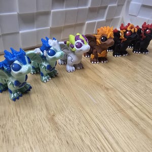 May include: A collection of colorful dragon figurines. The dragons vary in color, including blue, green, white, brown, orange, and black. Each figurine has detailed features, including wings and scales. They are arranged on a wooden surface.