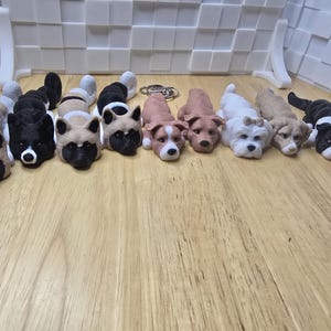 May include: A collection of dog-shaped figurines in various colors, including tan, black, and white. The figurines are lying on a wooden surface. Some have keychains attached.