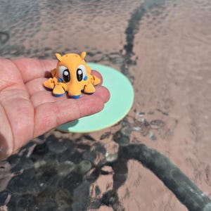 May include: A small, orange figurine with large black eyes and blue accents. The figurine has a cartoonish design, with rounded features and small appendages. It is held in a person's hand, with a light green circle in the background.
