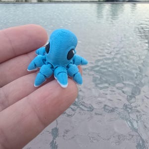 May include: A small, light blue octopus figurine with black eyes and white-tipped tentacles. The octopus is a 3D-printed design, showcasing a detailed, textured surface. The figurine is held in a hand, with a blurred background.