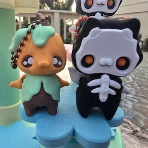 May include: Two whimsical figurines, one resembling a pumpkin with a green leaf-like top and the other a skeleton cat. The pumpkin figure is orange, brown, and green, while the cat is black and white. Both have large, expressive eyes and are on a blue flower-shaped base.