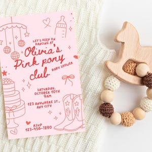 May include: A pink baby shower invitation with red text and illustrations, including a cake, cowboy boots, and a mobile. A wooden rocking horse and a beaded rattle with wooden and crocheted beads are also present.