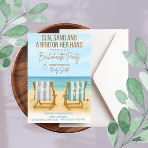 May include: Bachelorette party invitation with beach theme. The invitation features text "Sun, Sand and a Ring on Her Hand" and "Bachelorette Party". Two beach chairs are on the beach. The invitation is on a wooden tray with a white envelope and green leaves.