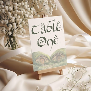 May include: A white card with green text that reads "Table One" in a whimsical font. The card is propped up on a small wooden easel. The background is a white fabric with a small bouquet of white flowers.