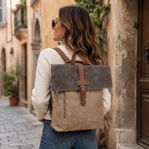 May include: A two-tone backpack featuring a tan lower section and a dark grey fold-over top flap. The bag includes brown leather-like straps, a central vertical buckle closure, a top handle, and a visible side zipper pocket on the tan portion.