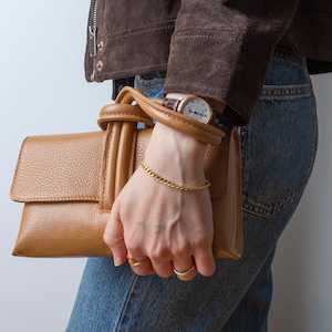 May include: A tan leather handbag with a top handle is held by a person wearing a gold bracelet and rings. A brown leather watch is on the person's wrist. The person is wearing blue jeans and a brown suede jacket.