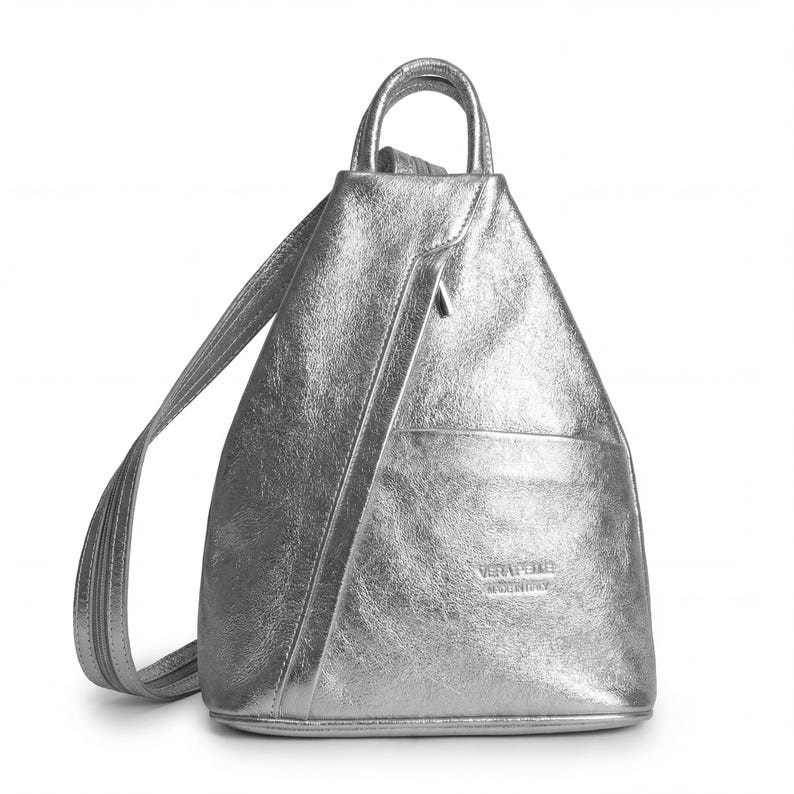 May include: Silver metallic backpack with a top handle and adjustable straps. Features a front pocket and a diagonal zipper. The bag has a textured finish and a triangular shape. The brand name is visible on the front.