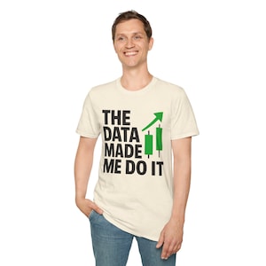 May include: A cream-coloured short-sleeve t-shirt featuring the black text THE DATA MADE ME DO IT alongside a green upward-pointing arrow and two green candlestick chart icons. The shirt has a crew neckline and a speckled fabric texture.