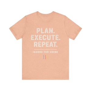 Op de afbeelding: Een perzikkleurig T-shirt met de witte tekst "PLAN. EXECUTE. REPEAT." en daaronder "IGNORE THE NOISE". Het ontwerp bevat drie verticale lijnen in rood, blauw en geel.