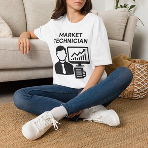 May include: A white short-sleeved t-shirt featuring a black graphic print of a person in a tie next to a computer monitor displaying an upward trending stock market graph and the text MARKET TECHNICIAN printed in bold black capital letters.
