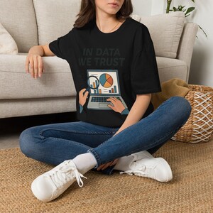 Può includere: Una t-shirt nera a maniche corte con la scritta IN DATA WE TRUST stampata in bianco sopra la grafica di un computer portatile che mostra un grafico a torta e a barre, con mani che tengono una lente d'ingrandimento sullo schermo.