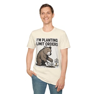 May include: A cream-colored short-sleeve t-shirt featuring a graphic of a brown bear digging in the dirt with a shovel. The text above the bear reads I'M PLANTING LIMIT ORDERS, and two small signs in the ground also read LIMIT ORDER.