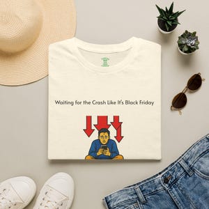 May include: A cream-colored t-shirt featuring the text "Waiting for the Crash Like It's Black Friday" above a graphic of a person looking at a smartphone with three red downward-pointing arrows positioned above their head.
