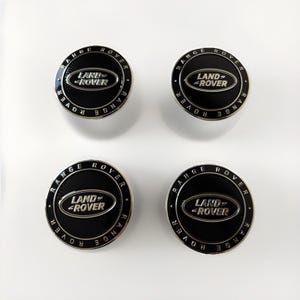 May include: Four black and silver Land Rover Range Rover wheel centre caps. Each cap features the Land Rover logo in silver against a black background, surrounded by the words "RANGE ROVER" in silver.