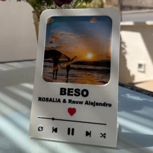 Może przedstawiać: Biały stojak na odtwarzacz muzyczny ze zdjęciem pary na plaży o zachodzie słońca. Stojak ma czerwone serce i napis "BESO ROSALIA & Rauw Alejandro".