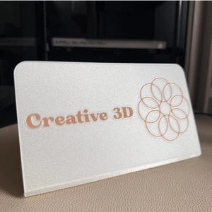Può includere: Un cartello bianco con il testo "Creative 3D" in lettere marroni e un design circolare marrone.