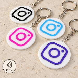 Può includere: Quattro portachiavi circolari bianchi con anelli e catene in metallo, ognuno con un logo Instagram di colore diverso: nero, rosa, azzurro e viola. I portachiavi sono dotati di tecnologia NFC, indicata dall'icona circolare NFC.