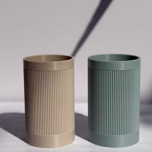 Puede incluir: Dos recipientes cilíndricos con detalles acanalados verticales. Uno es de color beige claro y el otro es verde azulado apagado. Los recipientes tienen un diseño simple y moderno, adecuados para guardar lápices, brochas de maquillaje o objetos pequeños.