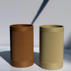 Può includere: Due contenitori cilindrici con dettagli a coste verticali. Uno è marrone caldo, l'altro verde oliva tenue. Entrambi hanno una base e un bordo pieni. I contenitori sono probabilmente destinati a contenere oggetti, come penne o pennelli per il trucco.