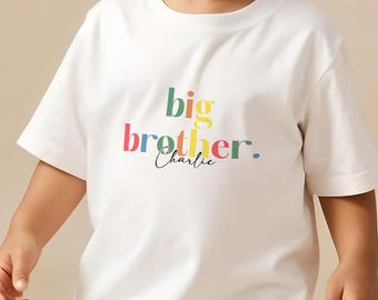 May include: A young child wearing a white t-shirt with the words "big brother" in colorful letters. The child is also wearing khaki pants and white sneakers. The shirt has the name "Charlie" printed below "brother". The brand name "Cubby & Coco" is in the top left corner.