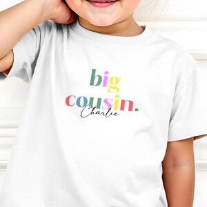 May include: A young child wearing a white t-shirt with the words "big cousin. Charlie" in colorful lettering. The child is also wearing blue shorts and smiling. The brand name "Cubby & Coco" is at the top left.