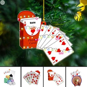 May include: A Christmas ornament featuring a deck of playing cards. The red card case is adorned with string lights and the words "Playing Cards." The cards display hearts and are strung with lights. The top card says "Name."