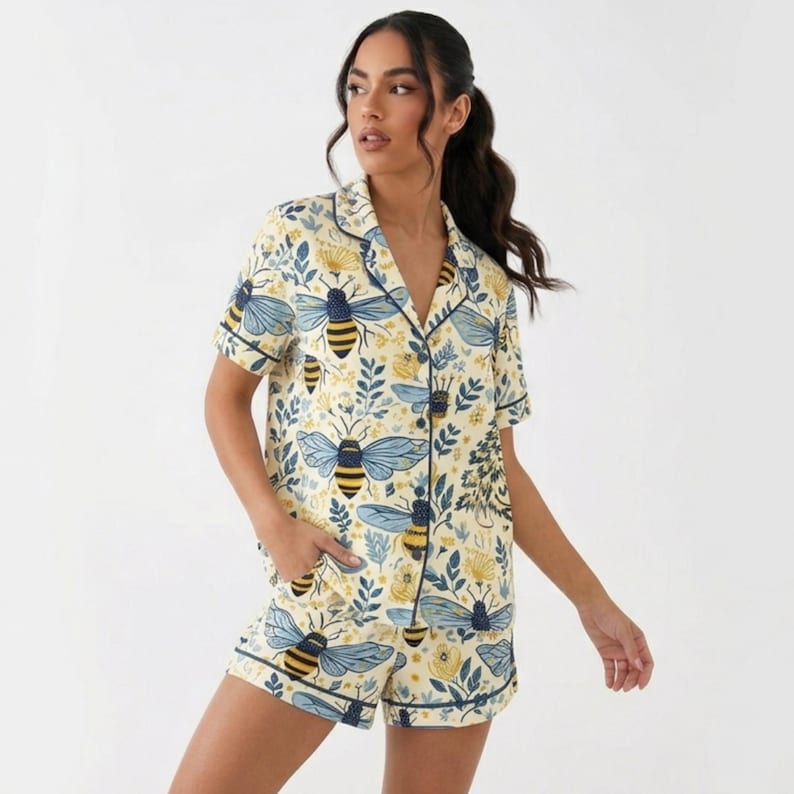 May include: A two-piece pajama set featuring a cream-colored background with a pattern of blue and yellow bees and floral elements. The set includes a short-sleeved button-down shirt and matching shorts, both trimmed with navy blue piping.