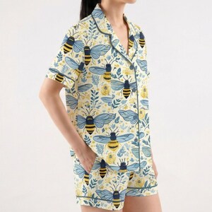 May include: A light yellow pajama set with a bee and floral pattern. The short-sleeved top and shorts feature a repeating design of blue and yellow bees, yellow flowers, and green leaves. The set has dark green trim.