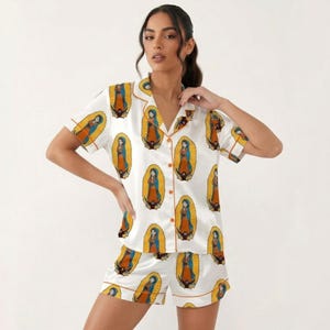 Pode incluir: Conjunto de pijama de cetim branco com uma ilustra&ccedil;&atilde;o repetida de uma figura religiosa em um halo dourado. A parte superior de manga curta e os shorts t&ecirc;m acabamentos laranja. O conjunto &eacute; feito de um material macio e sedoso.