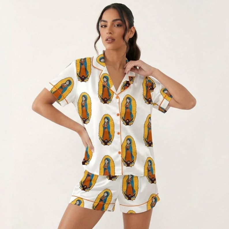 Pode incluir: Conjunto de pijama de cetim branco com uma ilustra&ccedil;&atilde;o repetida de uma figura religiosa em um halo dourado. A parte superior de manga curta e os shorts t&ecirc;m acabamentos laranja. O conjunto &eacute; feito de um material macio e sedoso.