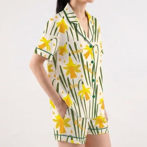 May include: A two-piece pajama set featuring a white background with a pattern of yellow daffodils and green stems. The set includes a short-sleeved button-down shirt and matching shorts. The words "Happy Mother's Day" are printed on the shirt.